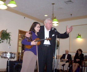 Kristin playing violin alongside an older gentleman speaking into a microphone